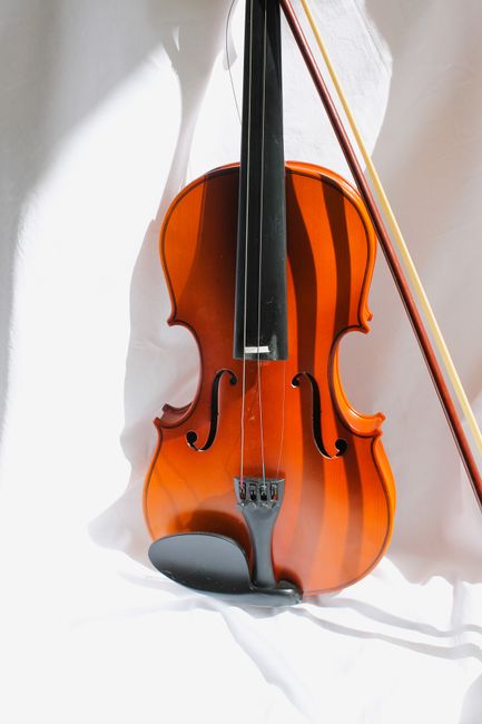 A close-up image of a polished wooden violin with a rich amber finish, positioned upright against a white fabric backdrop. The violin's strings, bridge, and ebony fingerboard are clearly visible, with the bow resting diagonally across the instrument, its horsehair and wooden stick extending beyond the frame. The scene suggests a focus on musical instruments, with no movement or personnel present. This image could relate to house removals involving the careful packing and transport of delicate items such as musical instruments, which Man and Van Bow may handle during furniture transport and home relocation services near Bow Road station, BOW.