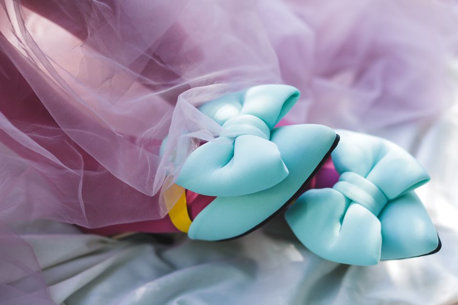 Close-up image showing a pair of light blue ballet flats with bow details on top, resting on a surface covered with soft, pastel-colored fabric and pink tulle material. The shoes are positioned near a transparent plastic wrapping, which is part of packing materials used during a home relocation or furniture transport process. In the background, there's a glimpse of cardboard boxes and packing supplies, indicating the ongoing packing and moving preparations. The scene is well-lit, emphasizing the textures of the fabric, shoes, and packaging materials, illustrating a typical stage in packing and loading procedures by a removals service like Man and Van Bow, assisting with household moves near Bow Road station.