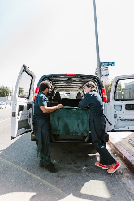 A man dressed in casual clothing is engaged in the loading process outside a residential property, working with several cardboard boxes of various sizes on the pavement beside a white delivery van wit