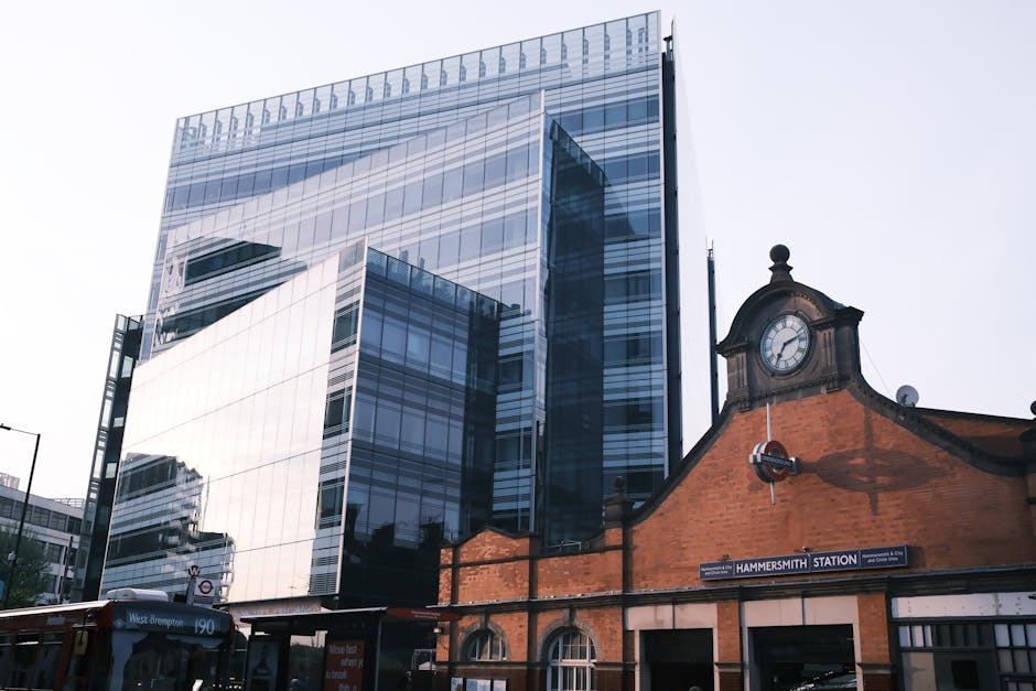A modern, multi-storey office building with a reflective glass facade occupying most of the image, standing behind an older, traditional brick structure with a clock tower and a sign for Hammersmith S